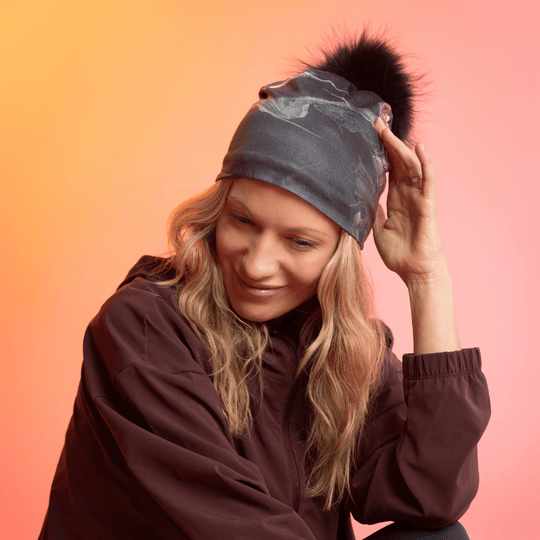 Femme portant une tuque gris foncé à motif tourbillonnant gris et un pompon noir, souriant sur fond dégradé.