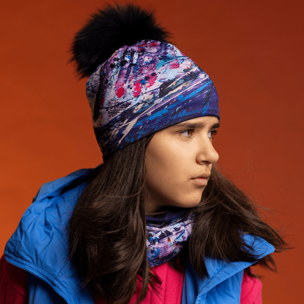 Jeune personne portant la Tuque cosmique avec pompon et un cache-cou assorti, avec une veste bleue, sur fond orange.