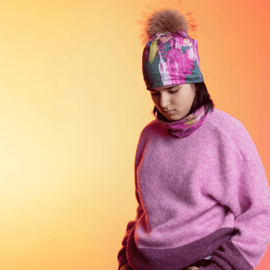 A young person wearing the Flower Glitch Beanie With Pompom and a matching neck gaiter with a fuzzy pink sweater, looking down against a warm gradient background.