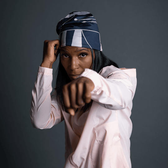 Une femme portant une tuque à rayures noires et bleues et un coupe-vent rose pâle, en position de boxe.