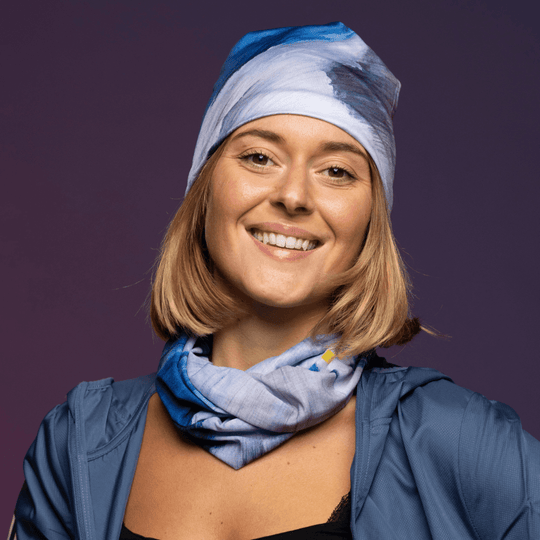 Une femme souriante porte une veste bleue et un bandana bleu comme cache-cou, avec un autre bandana noué en tuque.