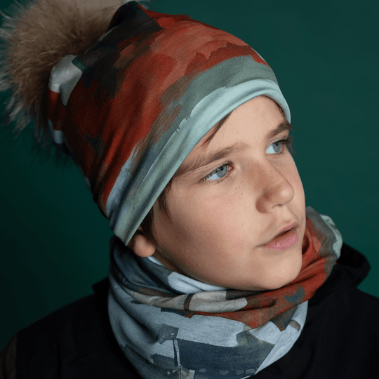 Une jeune personne porte une tuque et un tube multifonction Lachine assortis, regardant pensivement sur le côté sur un fond bleu sarcelle foncé.