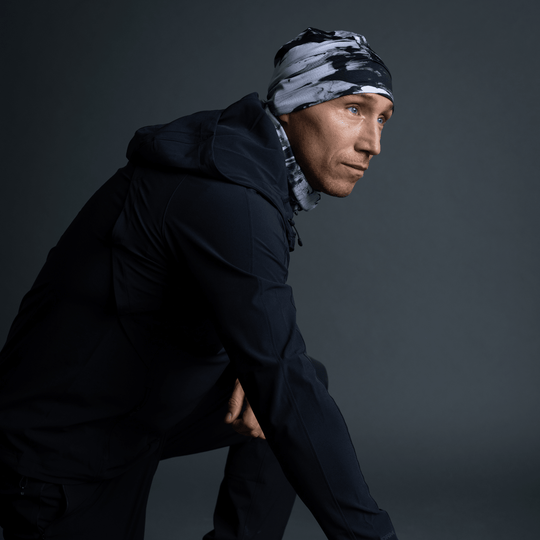 Homme en veste noire, tuque et cache-cou au motif abstrait noir et blanc, en pose accroupie.