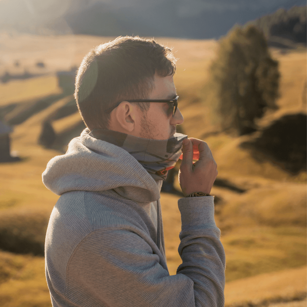 Un homme portant des lunettes de soleil et un sweat à capuche gris ajuste un cache-cou multifonction.