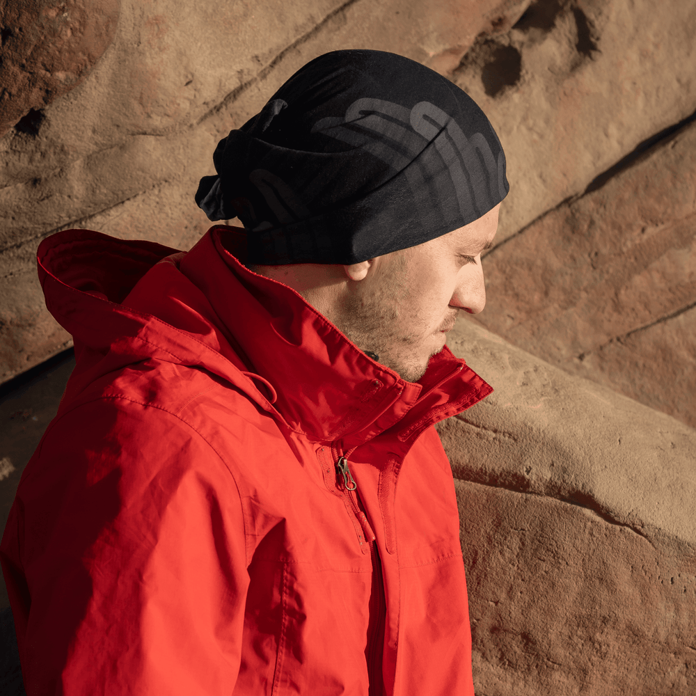 Un homme portant une veste rouge et un tube multifonction noir en bandana, debout devant un décor rocheux.