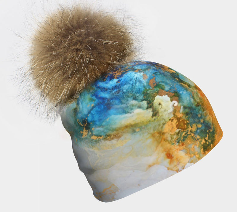 Close-up of a colorful 'Ocean Marble' print beanie with a large, fluffy natural fur pompom on a white background.
