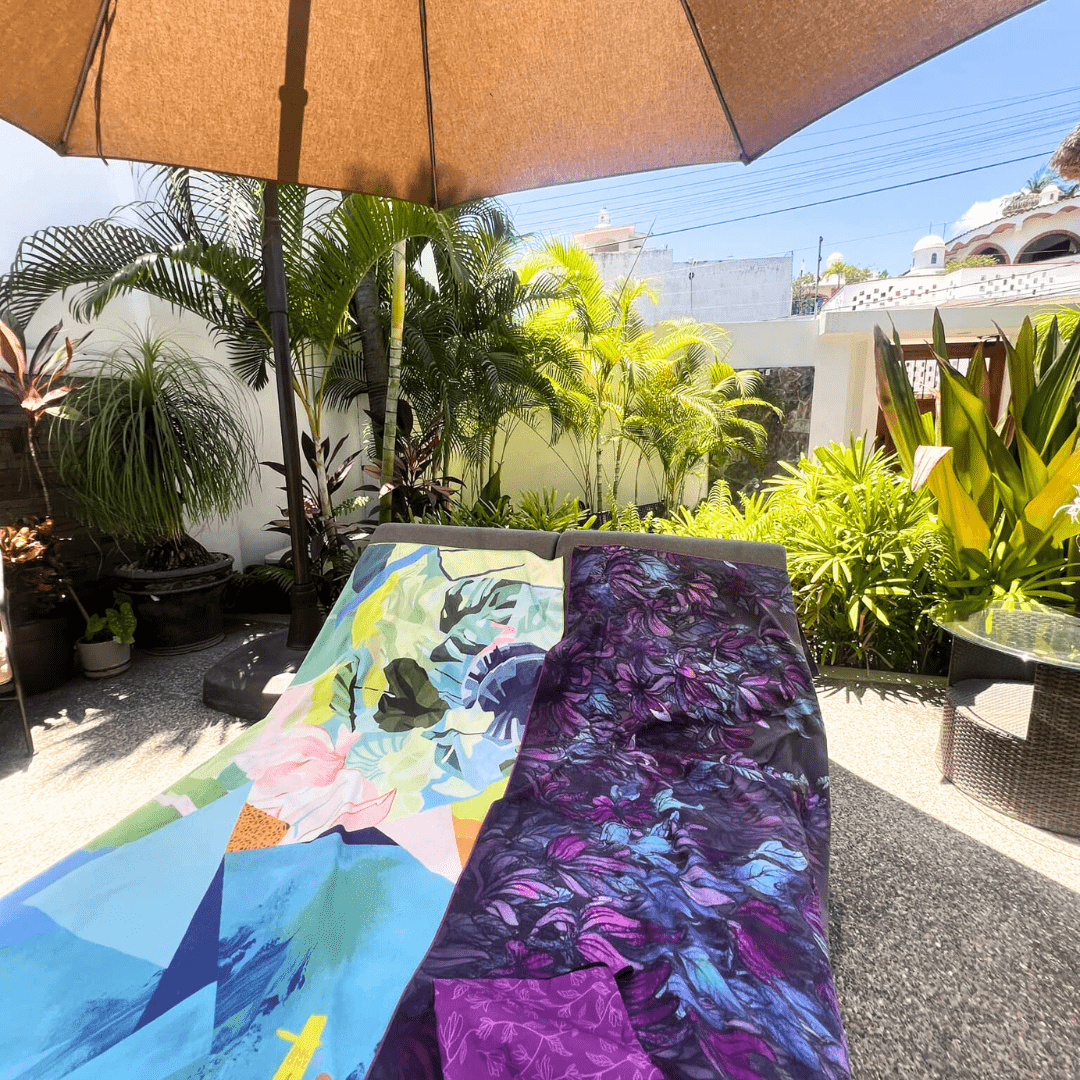 Two large microfiber towels, one with abstract floral art and the other with a purple floral design, spread out on lounge chairs under an umbrella.