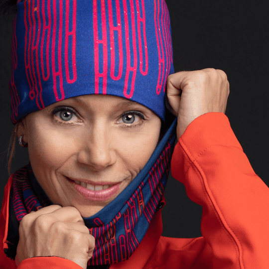 Une femme souriante, mettant une tuque à motifs bleus et rouges et montrant un cache-cou assorti, portant une veste orange.
