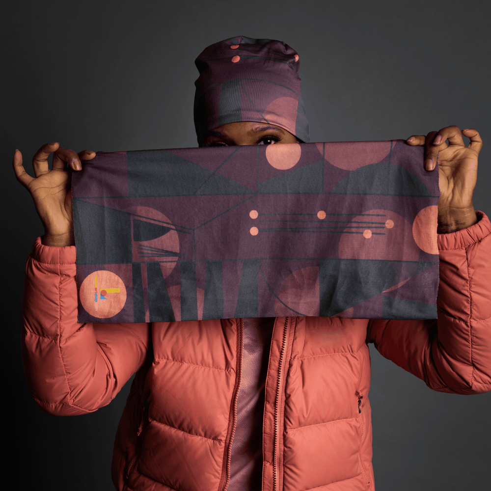 Une personne portant une veste matelassée corail tient un tube en tissu rectangulaire avec un motif géométrique violet et gris foncé, le visage partiellement masqué.
