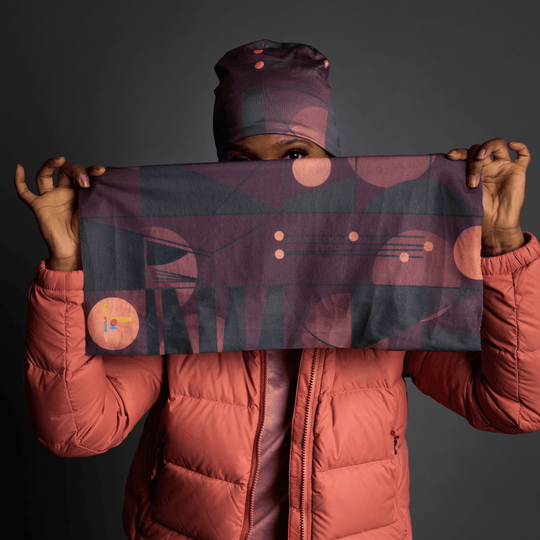 Une personne portant une veste matelassée corail tient un tube en tissu rectangulaire avec un motif géométrique violet et gris foncé, le visage partiellement masqué.
