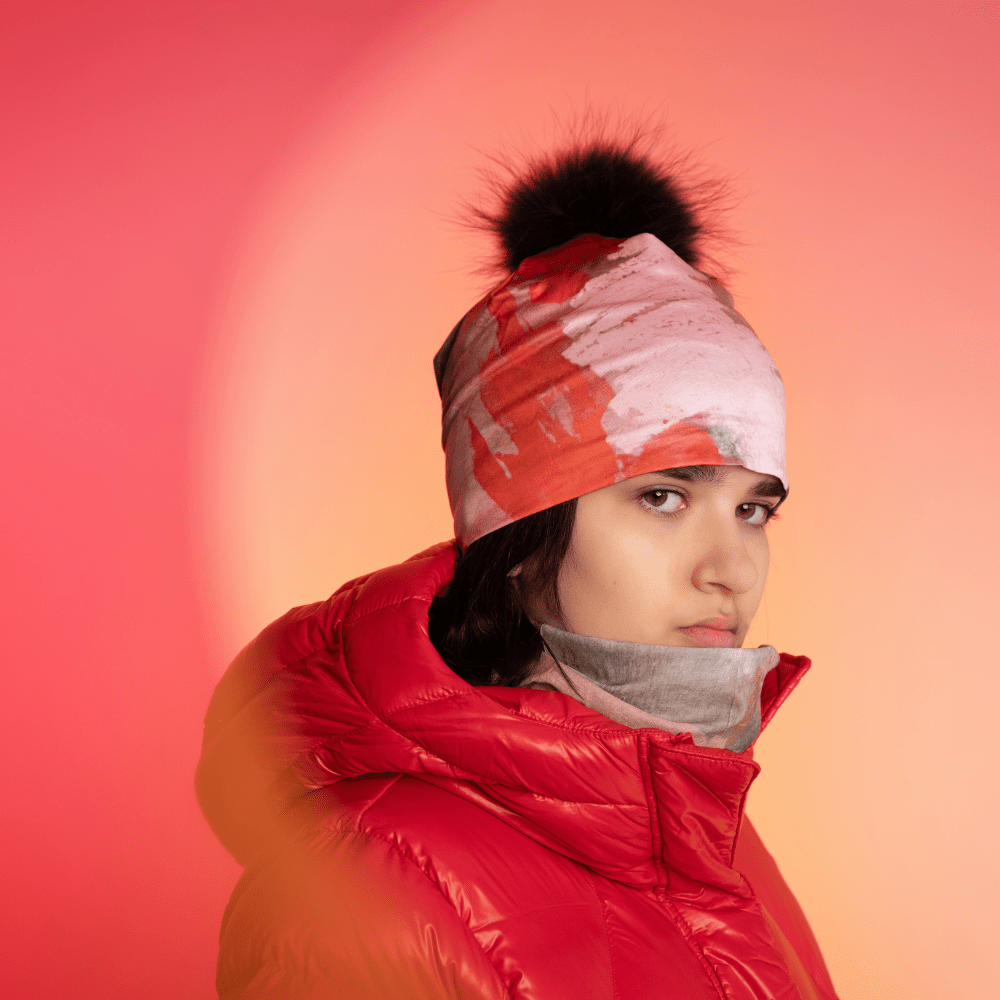 Young woman wearing a red puffer jacket, with the POLAR Pink &amp; Flames pattern as a beanie and a grey neck warmer, against a vibrant pink and orange gradient background.
