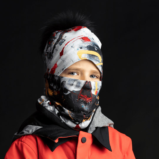 Un enfant portant le tube artistique rouge et noir comme masque facial et tuque, dans une tenue de ski.