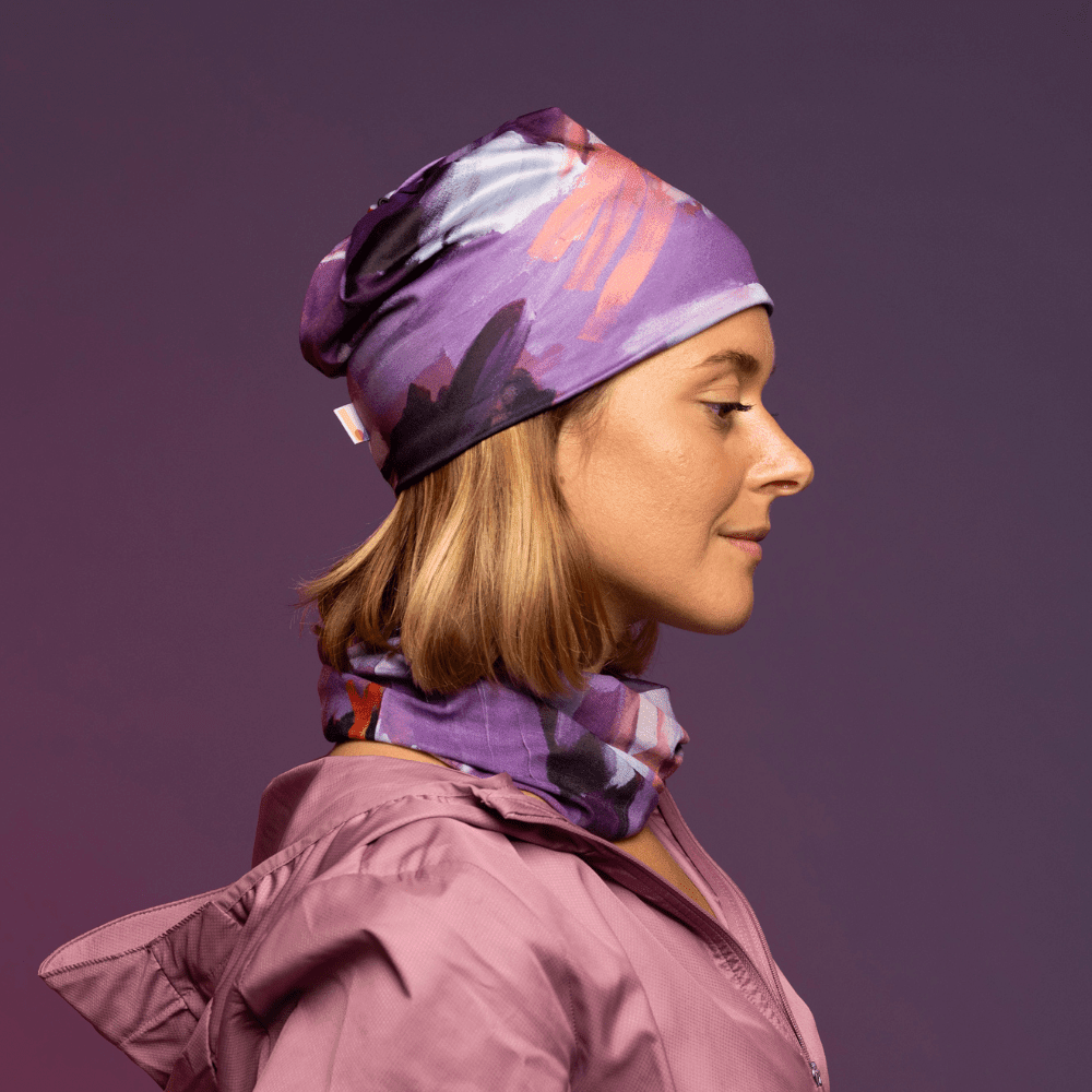 Une femme portant un tube et une tuque multifonction à imprimé abstrait violet, rose et blanc sur une veste rose.