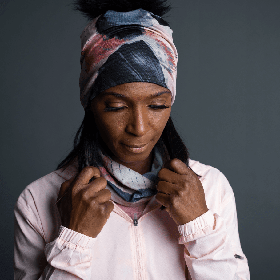Une femme portant une veste rose pâle, avec le tube multifonction POLAR Vie en Rose stylé en bandeau et cache-cou, révélant une tuque assortie avec un pompon.