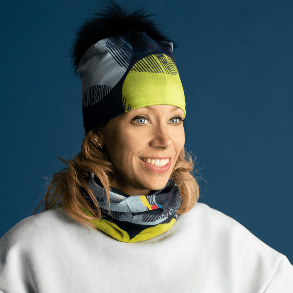 Une femme souriante portant une tuque et un cache-cou assortis à motif jaune-points, sur fond bleu.