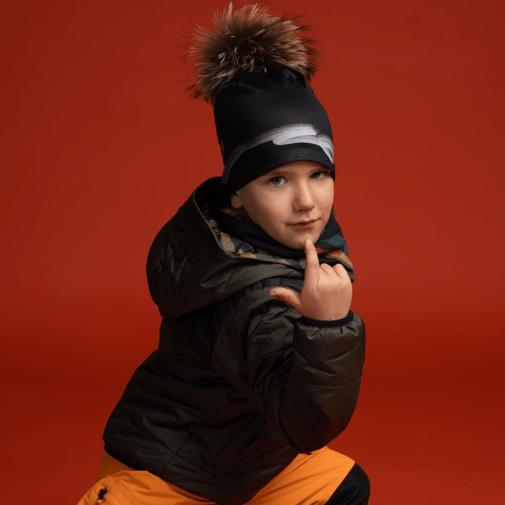 Un jeune garçon portant la Tuque Signature noire avec pompon et une veste d'hiver, posant devant un fond rouge.