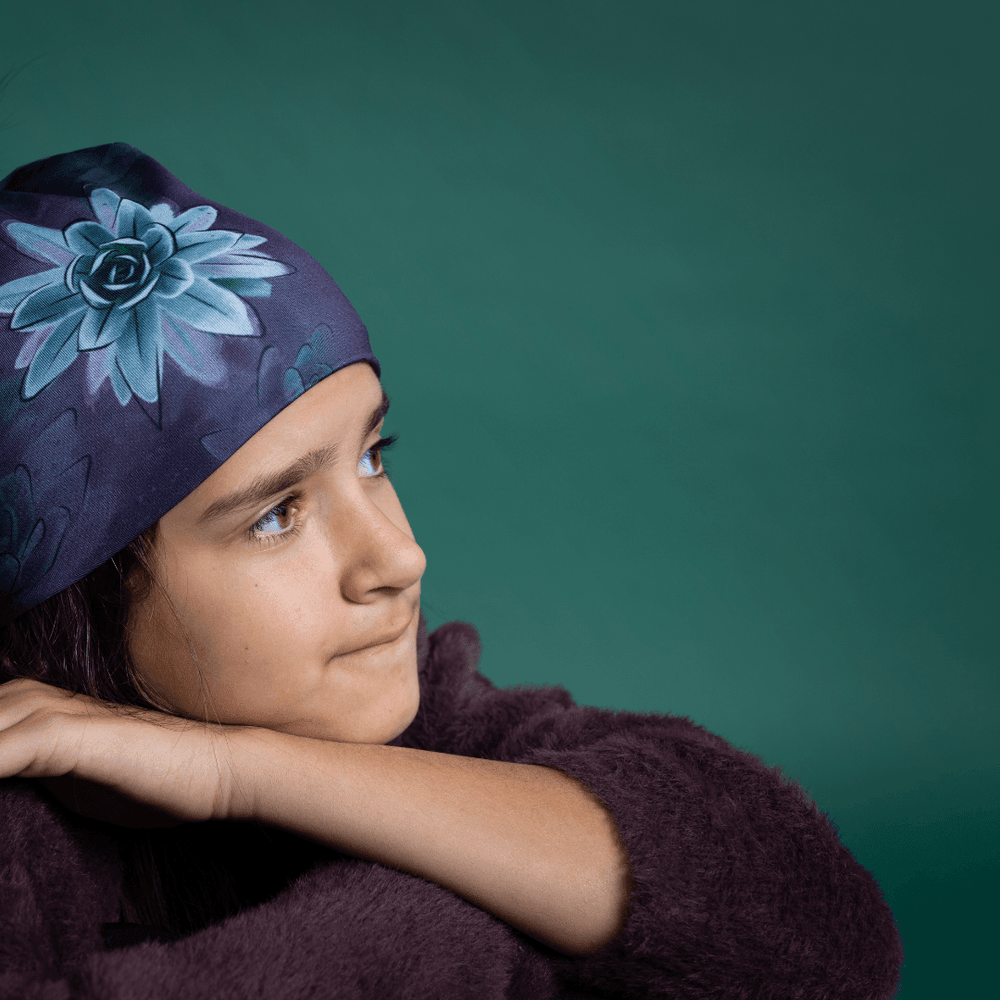 Jeune fille aux cheveux foncés portant la Tuque Succulente et un chandail duveteux violet foncé, regardant pensivement sur le côté sur fond sarcelle.