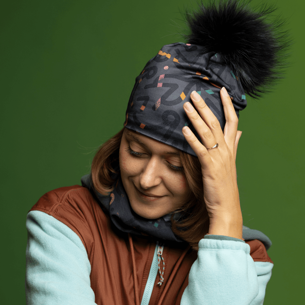 Femme portant une tuque grise avec pompon noir et cache-cou assorti, regardant pensivement vers le bas.