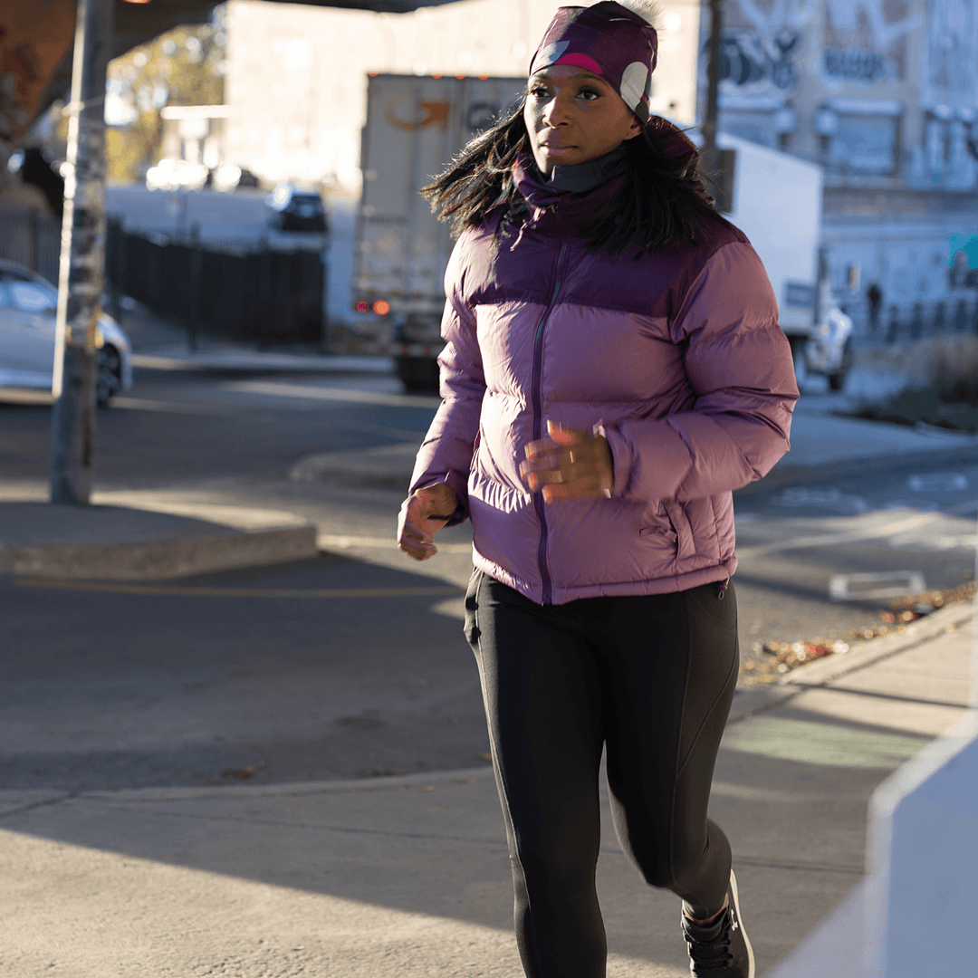 Woman running outdoors wearing a purple puffer jacket and a magenta beanie with a gray pompom.