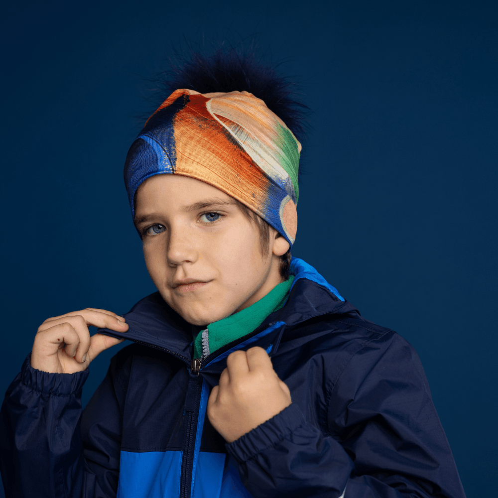 Jeune garçon portant la tuque jaune et bleue avec pompon en fausse fourrure marine amovible, stylisée avec une veste.